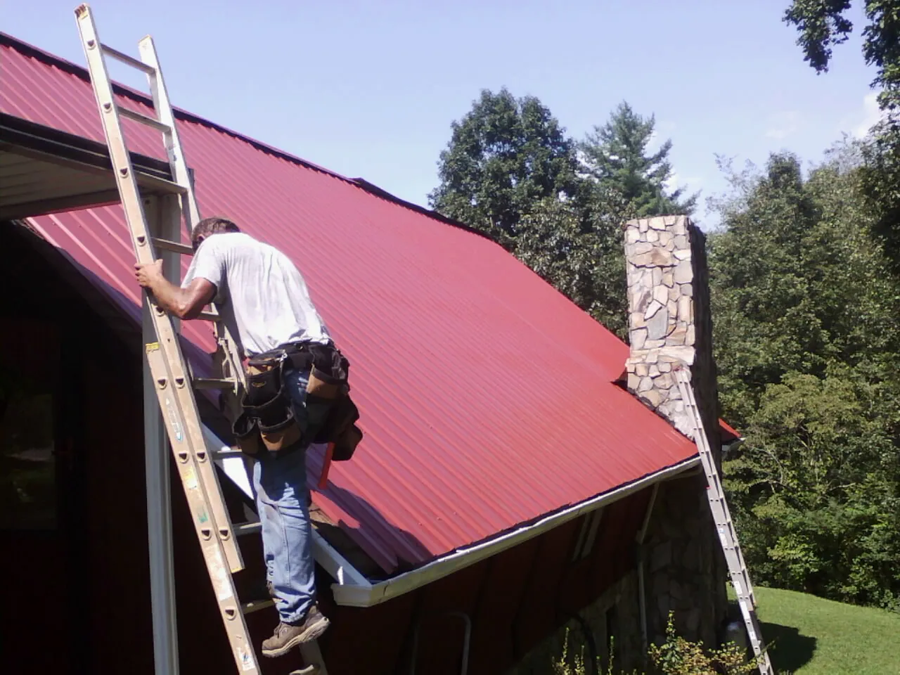 Roofer on a ladder beside a red metal roof and stone chimney — Soffit & Fascia Repair by Roofing Brothers in Forest Lake